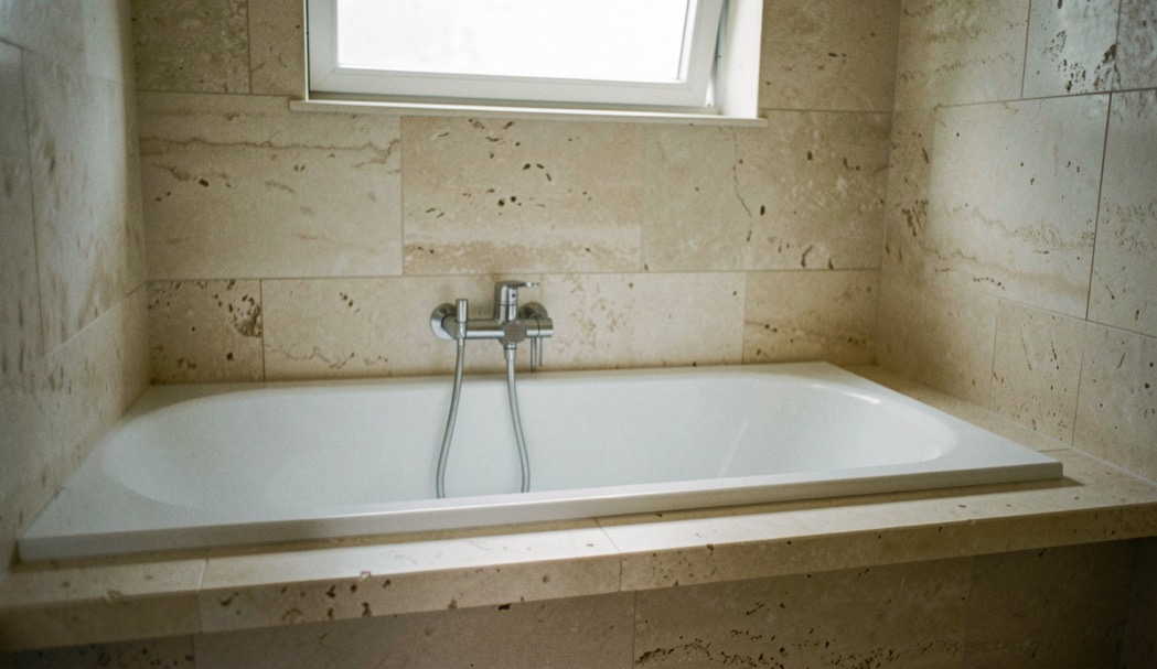 Bagno rivestito in lastre di travertino chiaro con vasca incassata e mensola in marmo sotto una luce naturale soffusa
